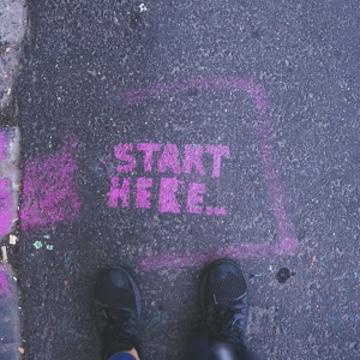 Two feet standing on a pavement in front of pink 'start here' graffiti